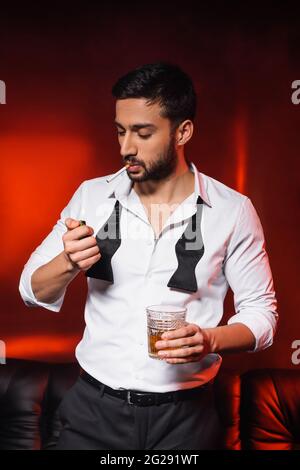 Homme barbu avec une cigarette et un whisky tenant un briquet sur fond noir avec un éclairage rouge Banque D'Images