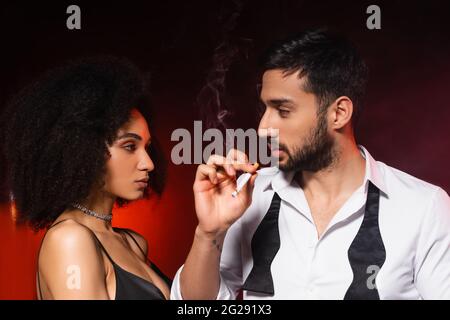 Homme fumant la cigarette près de l'élégante petite amie afro-américaine sur fond noir avec éclairage rouge Banque D'Images