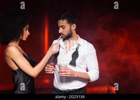 Femme afro-américaine tenant un briquet près d'un petit ami avec une cigarette et un whisky sur fond noir avec un éclairage rouge et de la fumée Banque D'Images