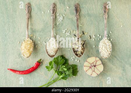 différents types de riz en cuillères avec épices. Riz sauvage, riz sushi, riz râpé et cuit à la vapeur sur fond de bois vert avec ail, piment et pars Banque D'Images