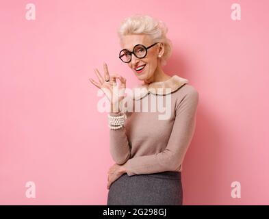 Grand-mère heureux dit bon travail ou bien fait, fait un geste correct. Photo de genre femme âgée porte des lunettes sur fond rose. Banque D'Images