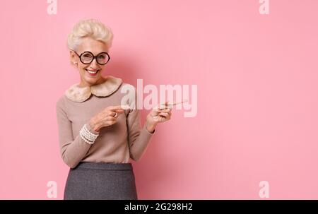 La femme souriante pointe à l'opposé des deux index, s'affiche sur l'espace de copie pour votre texte. Photo d'une femme âgée sur fond rose. Banque D'Images