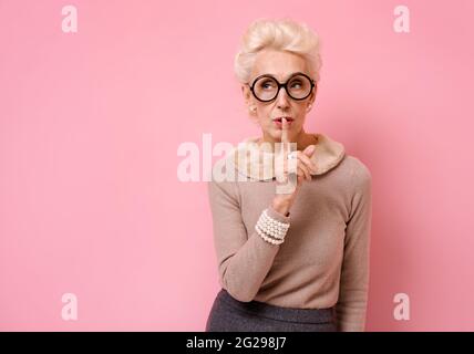 Gardez mon secret ! Belle grand-mère tenant le doigt sur les lèvres et montrant le geste de silence. Photo d'une femme âgée sur fond rose. Banque D'Images