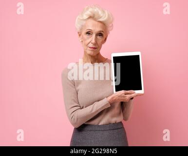 Femme souriante tenant une tablette numérique avec un écran vierge. Photo d'une femme âgée sur fond rose. Banque D'Images