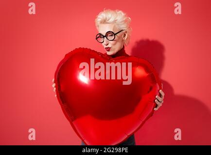 Une femme heureuse tient un ballon rouge en forme de coeur. Photo d'une femme âgée souriante amoureuse sur fond rouge. Saint Valentin Banque D'Images