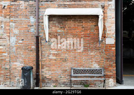 Devant un mur de briques abîmé, il y a un vieux banc et un collecteur de déchets sous un auvent blanc à côté d'une porte ouverte haute. Banque D'Images