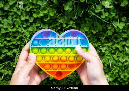 L'enfant joue sur l'herbe avec un jouet simple et relaxant. Pop il jouet en forme de coeur, couleur arc-en-ciel dans les mains des garçons le jour de l'été. Banque D'Images