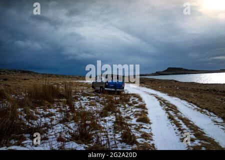 Véhicule bleu stationné près d'une route enneigée contre un ciel couvert gris sur la rive d'un lac calme, par temps froid d'hiver en Islande Banque D'Images