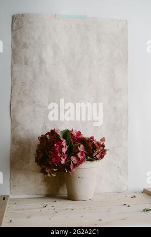 Fleurs d'Hydrangea rose aromatique dans un pot en argile grise sur une table en bois rustique sur une feuille de papier vierge pendue sur un mur dans une salle lumineuse Banque D'Images