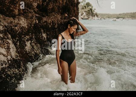 Bonne jeune femme ethnique en maillot de bain noir debout dans la mer à côté de la falaise et souriant Banque D'Images