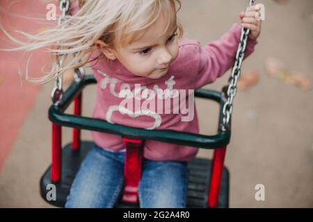 De dessus petite fille dans une tenue décontractée assis sur balançoire et regardant loin tout en passant du temps sur le terrain de jeu Banque D'Images