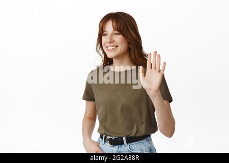 Image d'une jeune fille moderne qui agite la main, en disant bonjour, en levant la paume pour faire un geste élevé, en souriant et en regardant de côté amical, en rencontrant un ami, en se tenant contre Banque D'Images