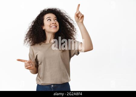 Bonne jeune femme choisissant entre deux produits, pointant latéralement et regardant vers le haut avec le visage satisfait, cliquez sur quelque chose, debout sur blanc Banque D'Images