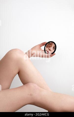 Rognez la jeune femme avec le symbole de naissance de nevus sous les yeux réfléchissant dans un petit miroir rond tout en regardant l'appareil photo sur fond blanc Banque D'Images