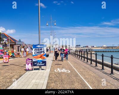 15 juin 2019: Lowestoft, Suffolk, Royaume-Uni - jeunes couples se promenant sur la promenade lors d'une journée d'été ensoleillée au bord de la mer, panneau pour les poissons et les frites. Banque D'Images