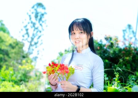 Les écolières en robe longue traditionnelle ou uniforme Ao Dai posant avec des fleurs phoenix école yard Mark les étudiants restent intemporels à long an, Vietnam Banque D'Images