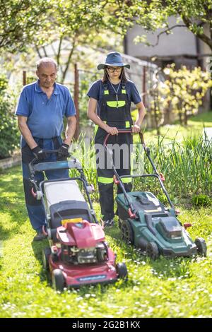 Grand-père âgé enseignant à sa petite-fille comment utiliser la tondeuse et couper l'herbe. Ils travaillent dans le jardin rustique du village. Banque D'Images