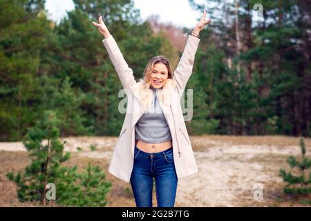 Jeune belle blonde longue cheveux femme debout vue de face avec les mains levées sur fond vert naturel extérieur. Jeune femme bras élevés appréciant Banque D'Images