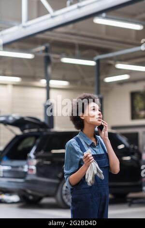 un jeune mécanicien afro-américain rêveur en combinaisons parlant sur smartphone en service de réparation automobile Banque D'Images