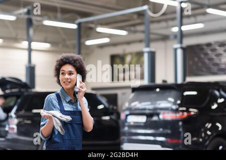 inquiet jeune mécanicien afro-américain dans des salopettes parlant sur smartphone dans le service de réparation automobile Banque D'Images