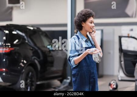 souriant jeune mécanicien afro-américain dans des salopettes parlant sur smartphone en service de réparation automobile Banque D'Images