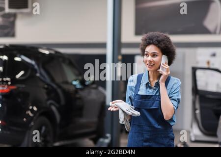 joyeux jeune mécanicien afro-américain en combinaisons parlant sur smartphone en service de réparation automobile Banque D'Images