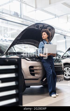 jeune mécanicien afro-américain debout avec un ordinateur portable près de voitures avec capot ouvert en service de réparation automobile Banque D'Images