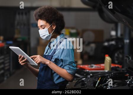 jeune mécanicien afro-américain en dactylographie de masque de protection sur tablette numérique près de la voiture dans le garage Banque D'Images