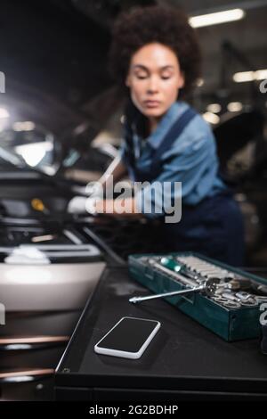 vue rapprochée du téléphone portable et clé dans la boîte à outils avec une femme afro-américaine floue travaillant dans le garage Banque D'Images