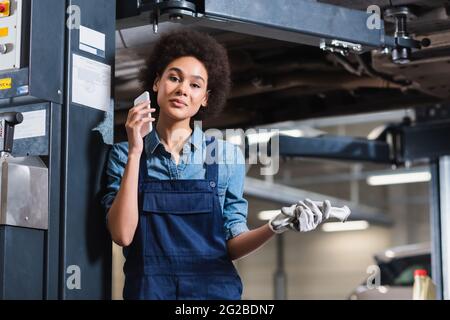 jeune mécanicien afro-américain souriant parlant au téléphone portable dans un garage Banque D'Images