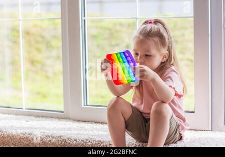 Petite fille blonde enfants jouer avec nouveau jouet sensoriel tendance - pop arc-en-ciel. Jouet antistress pour les enfants et les adultes.jouet coloré simple bosseler. Squis Banque D'Images