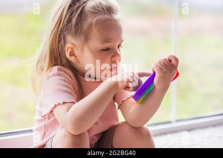 Une petite fille blonde s'assoit près de la fenêtre et joue avec un nouveau jouet sensoriel tendance - pop arc-en-ciel. Antistress jouet très simple fossle. Doux et accroupi b Banque D'Images