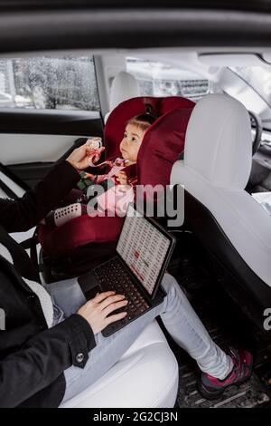 Bébé fille assis dans le siège de voiture pendant que la mère utilise un ordinateur portable Banque D'Images