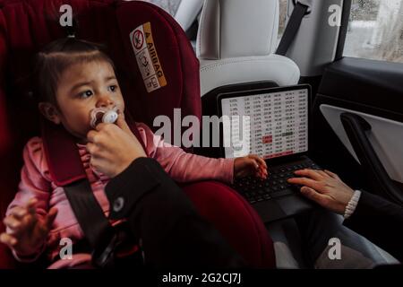 Bébé fille assis dans le siège de voiture pendant que la mère utilise un ordinateur portable Banque D'Images