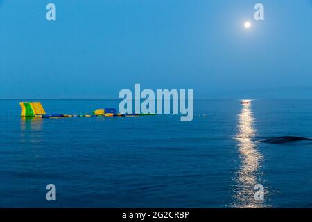 Clair de lune sur la mer, jeu gonflable jaune, bateau dans le reflet de la lumière sur l'eau. Moriani-Plage, Corse, France Banque D'Images