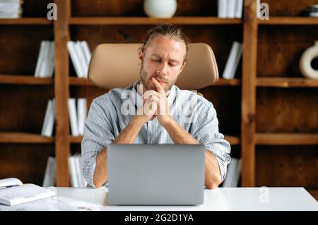 Pensive focalisé sérieux jeune adulte caucasien homme, pdg ou employé de bureau, assis dans le bureau à la table, à l'aide d'un ordinateur portable, reposant sa tête sur ses mains pense et analyse de la stratégie d'entreprise Banque D'Images