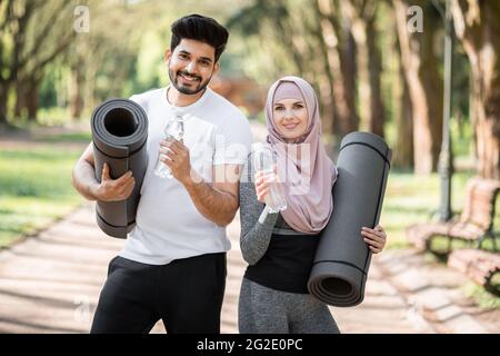 Couple musulman actif en vêtements de sport posant un parc vert avec une bouteille d'eau et un tapis de yoga entre les mains. Concept de rafraîchissement, de loisirs et de soins de santé. Banque D'Images