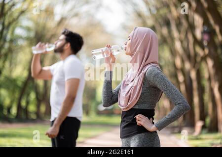 Vue latérale de la femme musulmane et de l'homme en activité en buvant de l'eau fraîche de la bouteille tout en se tenant au parc de la ville. Jeune couple prenant une pause pendant l'exercice matinal à l'extérieur. Banque D'Images