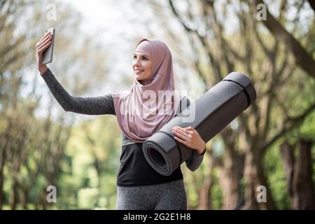 Bonne femme musulmane dans une tenue de sport et hijab emportant le selfie sur un smartphone moderne tout en se tenant au parc vert avec tapis de yoga a la main. Concept