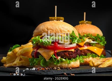 Deux grands hamburgers juteux avec du bœuf, du fromage, des légumes et des frites sur fond noir. Vue latérale, gros plan. Banque D'Images