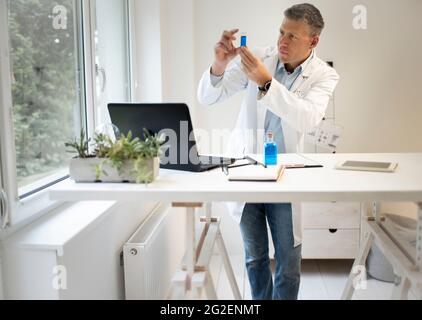 homme chimiste, scientifique travaille à la table debout et examine les liquides bleus dans de petits flacons et porte un manteau blanc Banque D'Images