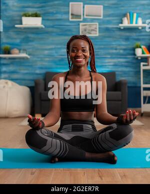Portrait d'une femme noire souriante pratiquant le yoga à la maison assise dans le lotus poser sur le tapis méditant, pratiquant la méditation de pleine conscience harmonie, africain américain faisant l'entraînement relaxant. Banque D'Images