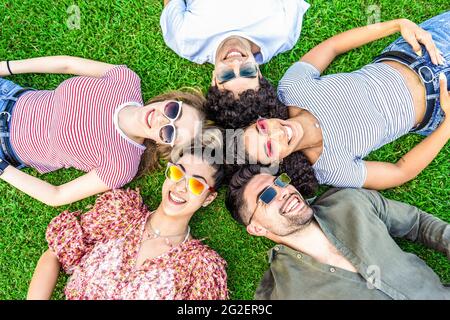 Groupe d'amis allongé sur le parc de prairie portant des lunettes de soleil colorées à la mode regardant la caméra rire. Heureux de divers internationaux appréciant des amis Banque D'Images