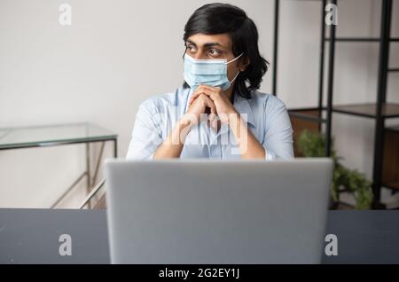 Un jeune Indien pensif aux cheveux foncés portant un masque médical de protection à l'aide d'un ordinateur portable assis au bureau, un employé de sexe masculin de l'est s'en prend bien à l'avenir Banque D'Images