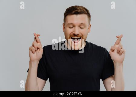 Beau jeune homme excité en t-shirt noir croisant les doigts et espérant la bonne chance Banque D'Images