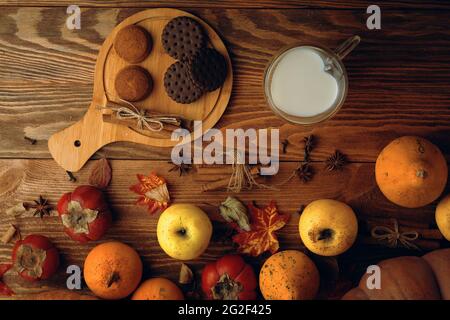 Biscuits au chocolat, légumes, fruits et lait dans une tasse en forme de coeur. Cuisson avec des bâtonnets de cannelle sur fond en bois. Composition d'automne. Pommes et persimmons, récolte d'automne. Banque D'Images