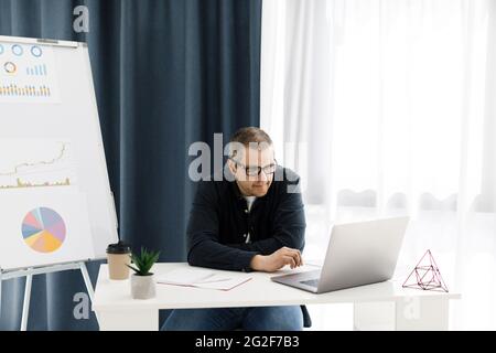 Beau gars en tenue décontractée à la maison, il tape sur le clavier et regarde sur l'écran. Navigation sur le Web, développement, freelance Banque D'Images