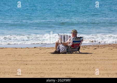 Homme assis lisant le quotidien sur la plage par une chaude journée ensoleillée à Bournemouth, Dorset, Royaume-Uni, en juin Banque D'Images