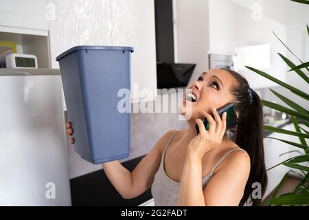 Worried Woman Holding seau tandis que les gouttelettes d'eau Fuite au niveau du plafond dans la cuisine Banque D'Images