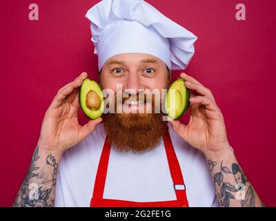 un chef heureux avec une barbe et un tablier rouge tient un avocat Banque D'Images